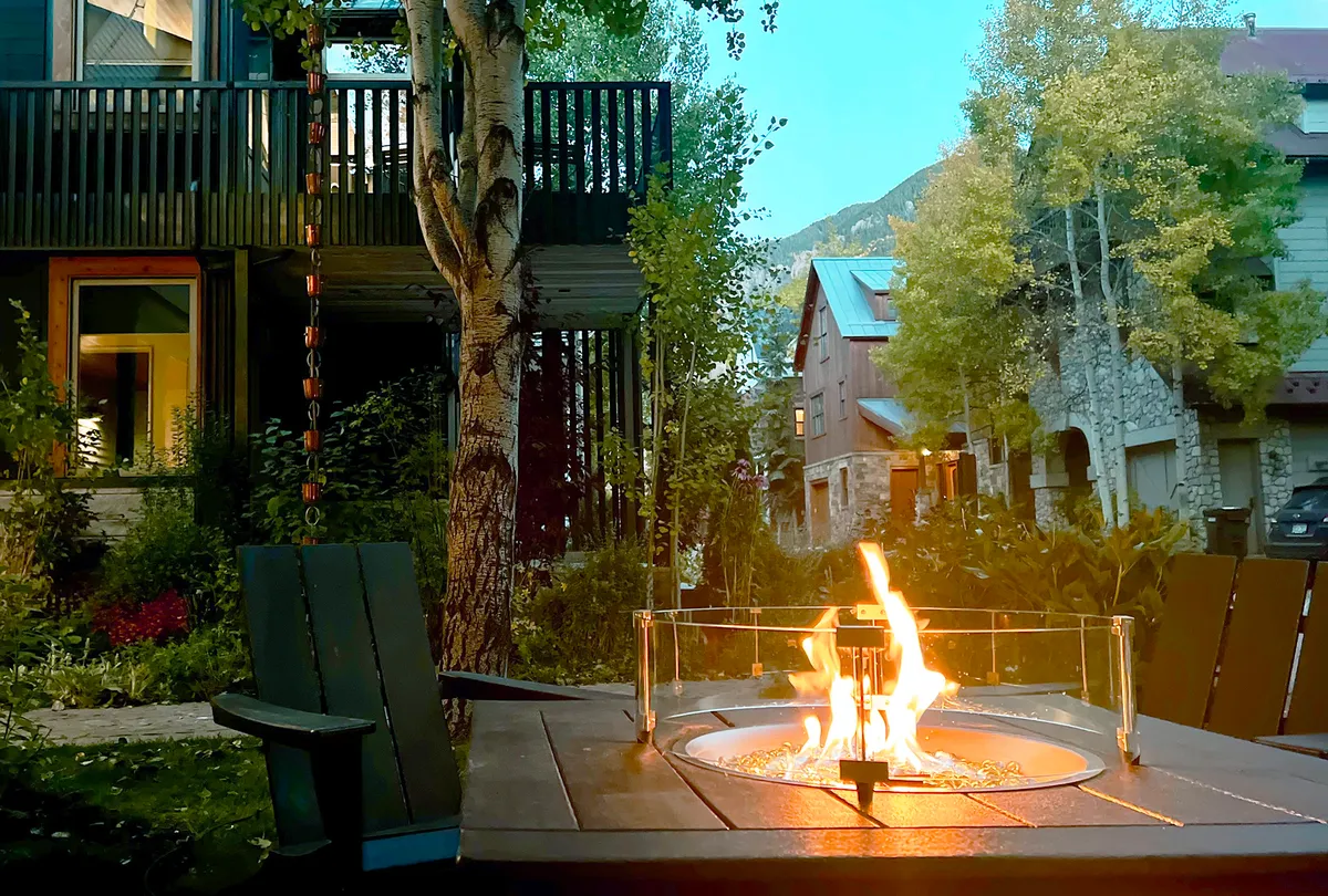 This exterior image captures a cozy fire pit on a deck surrounded by trees, with a stunning mountain view in the background. The scene evokes warmth, relaxation, and the joys of gathering outdoors, no matter the season.