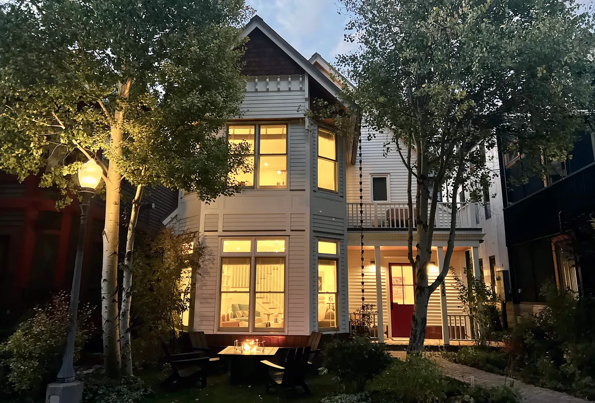An evening exterior image showing illuminated windows of the house, a welcoming outdoor seating arrangement, trees surrounding the property, and a pathway leading to the entrance. The ambiance is inviting and signals a space ideal for family gatherings both inside and out.