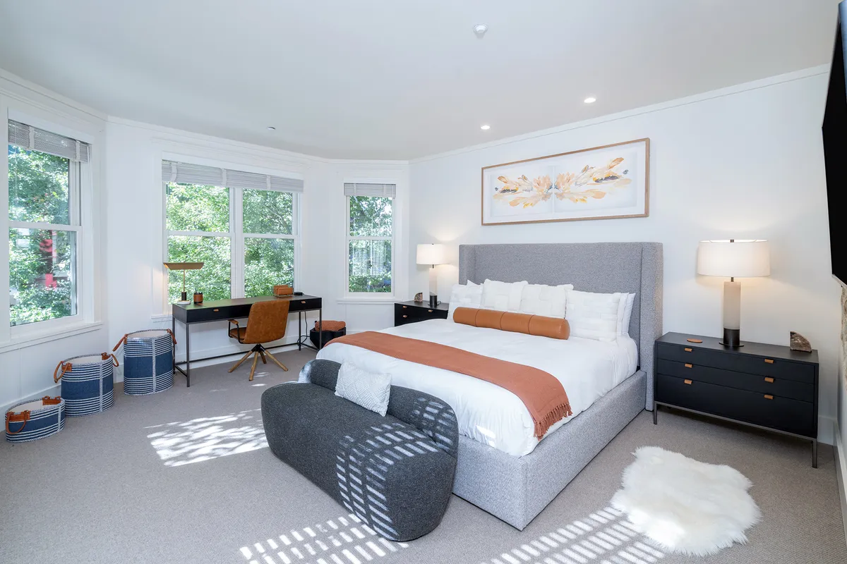 A serene bedroom featuring a California king bed adorned with soft decorative pillows, a desk with a chair for remote work, large windows letting in natural light, tasteful artwork, and a plush rug. The overall décor is calming and uncluttered, emphasizing comfort and relaxation.
