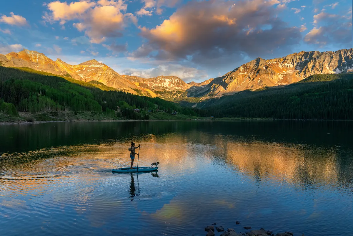 A paddleboarder glides across calm water, accompanied by a dog, with majestic mountain reflections and dramatic clouds in the background. The tranquil lake is surrounded by lush greenery, evoking a sense of outdoor adventure and natural beauty.