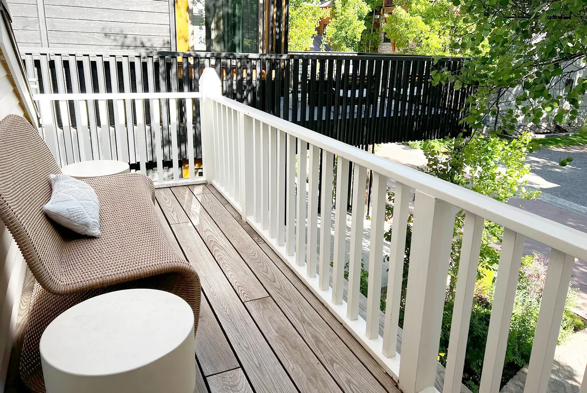 A tranquil outdoor seating area with a rattan-style chair, white side tables, and green foliage, set against the backdrop of a peaceful residential street. The space feels inviting for a relaxing moment after a day out.