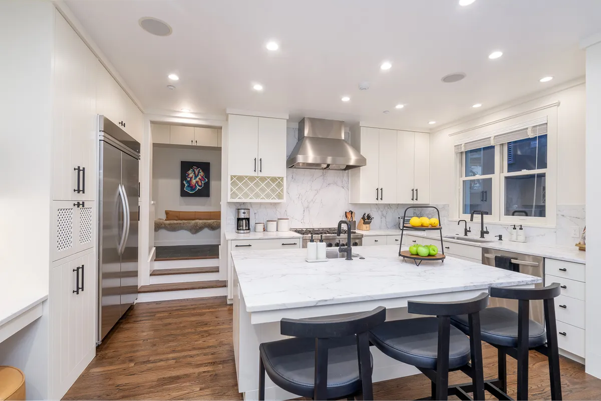 A modern gourmet kitchen with a large island, gleaming stainless steel appliances, marble countertops, and stylish black bar stools. A colorful fruit display adds a touch of homey warmth, and the space feels both functional and ideal for entertaining.