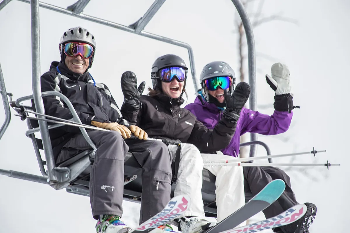 Three skiers are seen riding a chairlift above a snow-covered landscape, with their ski equipment visible. The scene is energetic and immersive, capturing the thrill of a day on the slopes and the close proximity to skiing activities.
