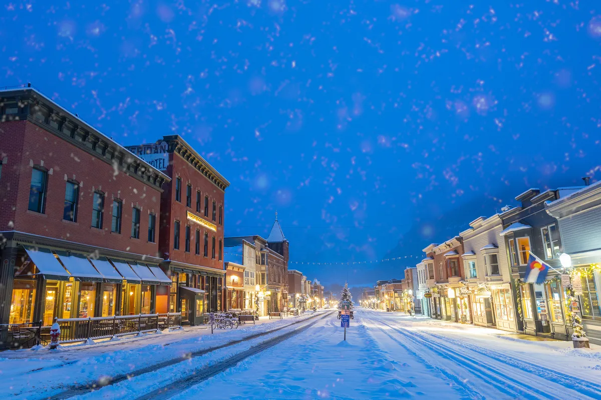 A picturesque snow-covered street lined with historic buildings, festive street lights, and a large decorated Christmas tree in the town center. The ambiance is lively, with people strolling and window shopping, evoking a sense of community and holiday charm.