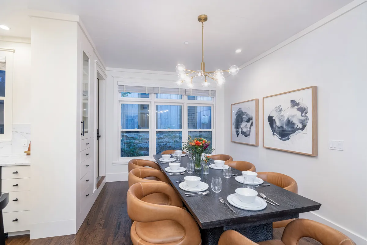 A beautifully set dining table for eight sits under a striking chandelier, with artwork on the walls and a window providing natural light. The table is ready for both intimate breakfasts and celebratory dinners, seamlessly integrated into the open living space.