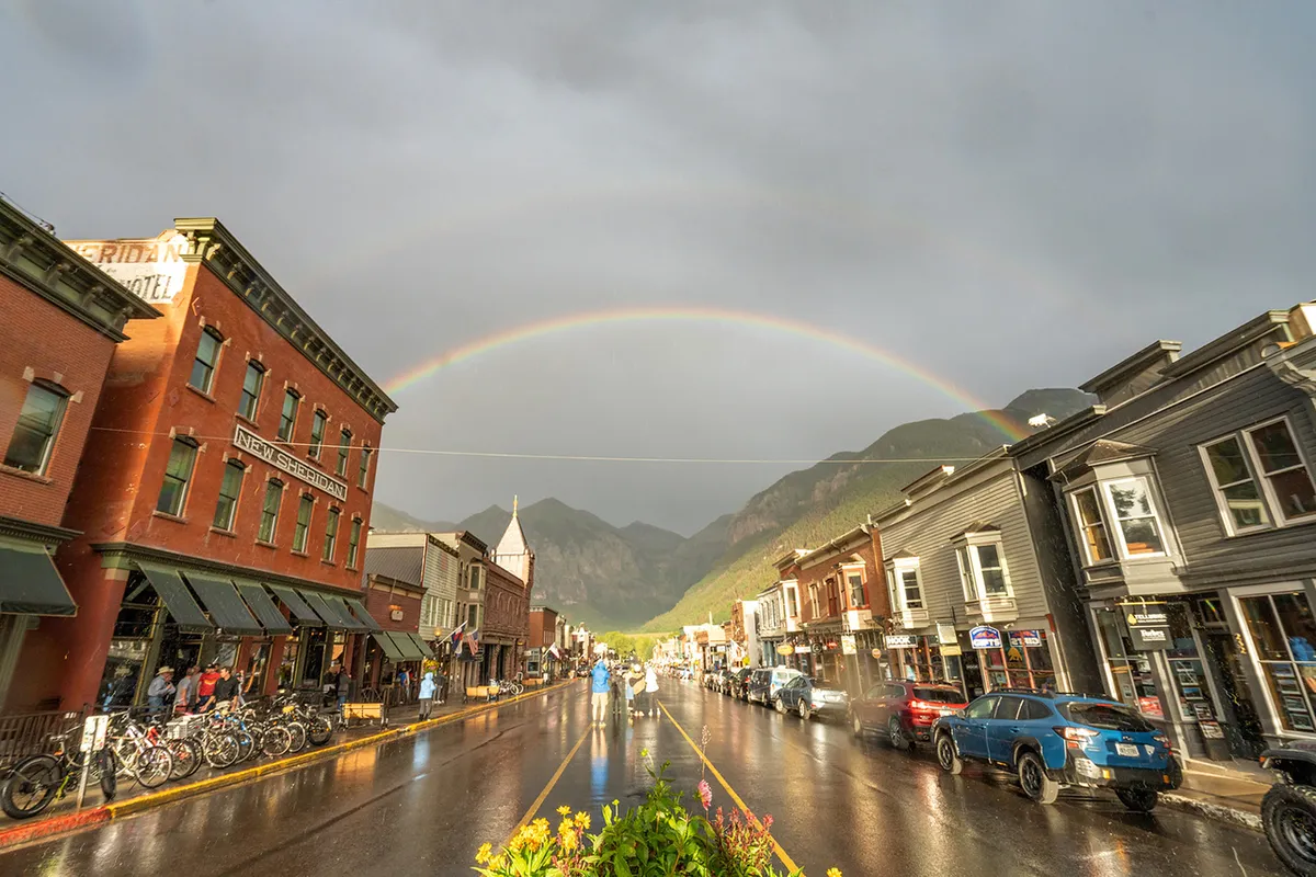 A snow-covered street lined with historic buildings and a vibrant rainbow arching above, set against a dramatic mountain backdrop. The wet street and festive atmosphere capture the lively spirit of Telluride’s downtown and its proximity to mountain adventures.