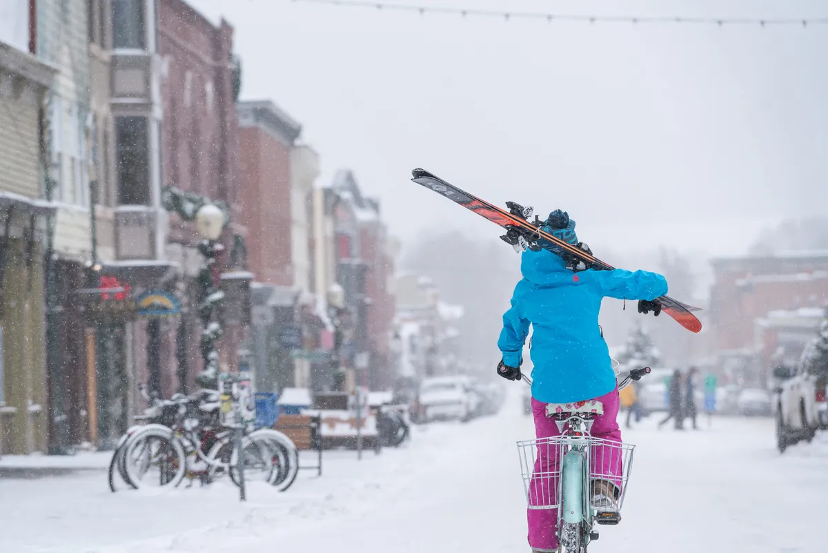 A lively winter street scene featuring a person riding a bike with skis on their shoulders, navigating a snow-covered street flanked by historic buildings. The mood is playful, capturing the intersection of adventure and local life.
