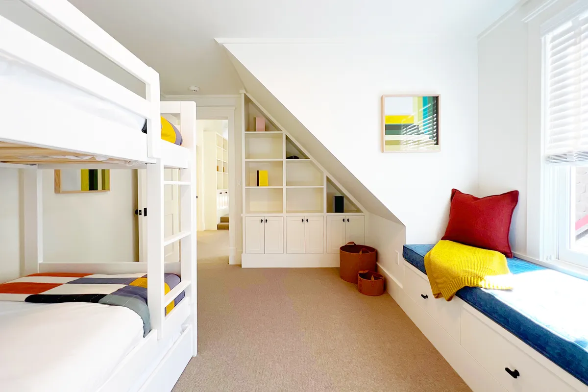 A bunk bedroom featuring built-in shelving, a window seat for reading, decorative wall art, and inviting bedding. The space looks comfortable and flexible, suitable for both relaxation and play, with room for trundles or extra guests.