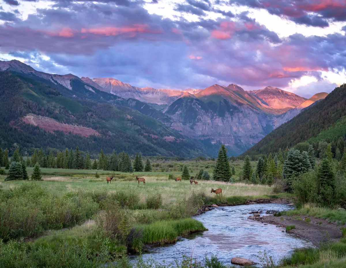 A scenic landscape featuring mountains, a flowing stream, and deer grazing nearby. Wispy clouds drift overhead, and the natural setting is peaceful yet full of opportunity for hiking and wildlife observation.